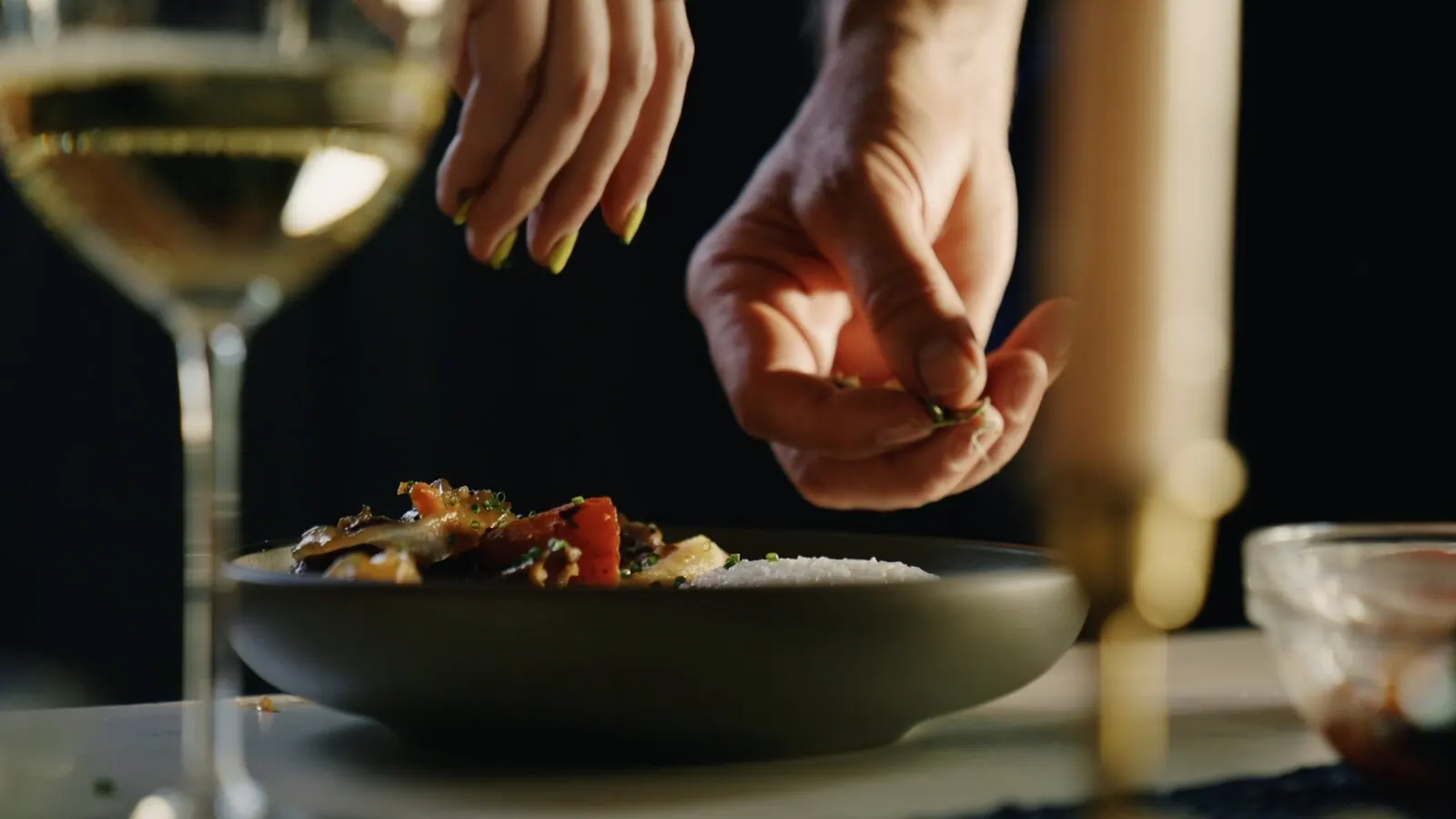 Hands finishing a plated stir-fry with fresh herbs next to a glass of white wine